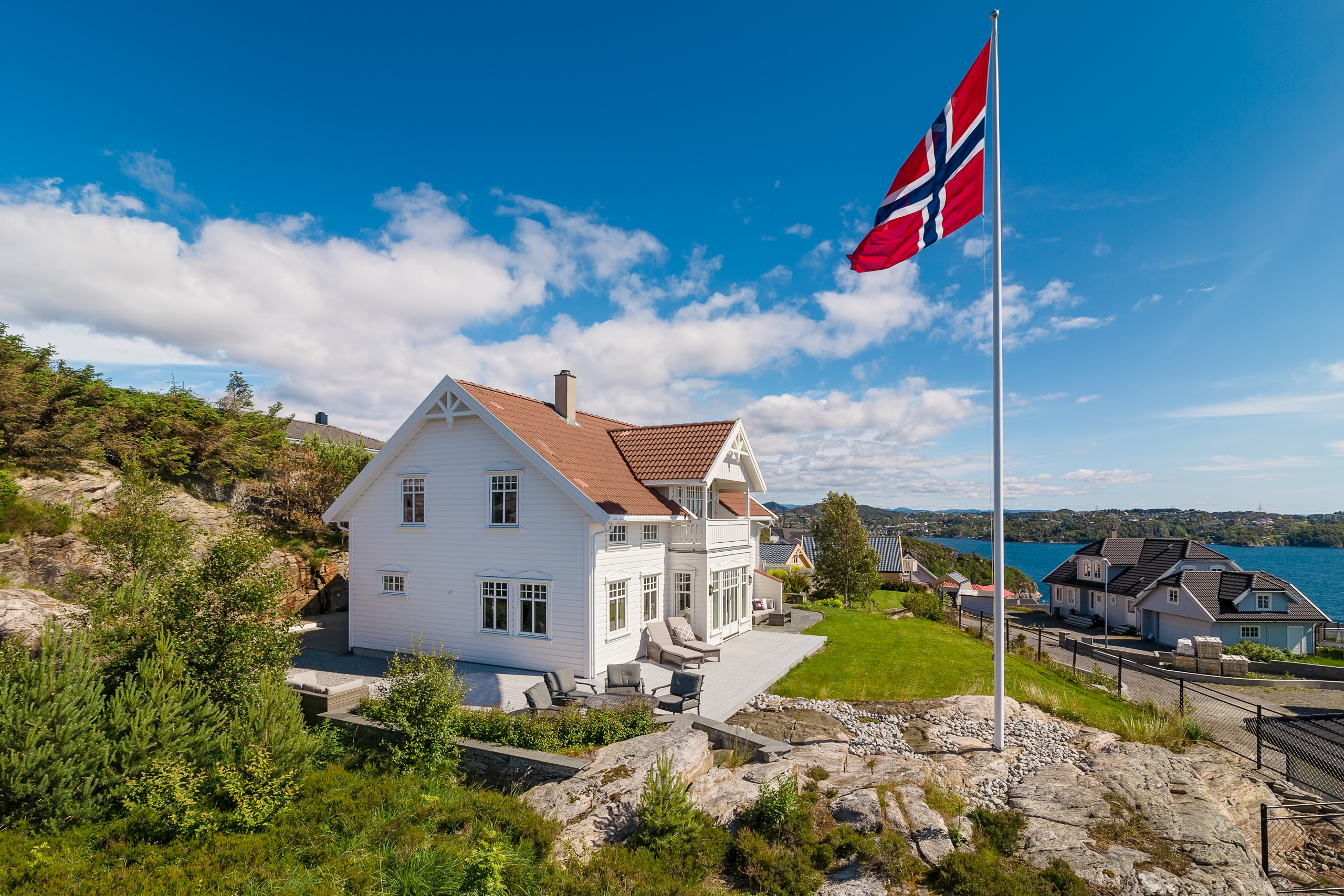 Mastfoto på en sommerdag av enebolig på stor tomt, det norske flagget i forgrunn.